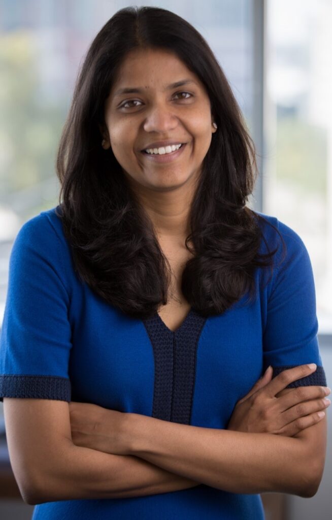 Archana Vemulapalli faces the camera with her arms crossed, smiling, in a blue short-sleeved top.