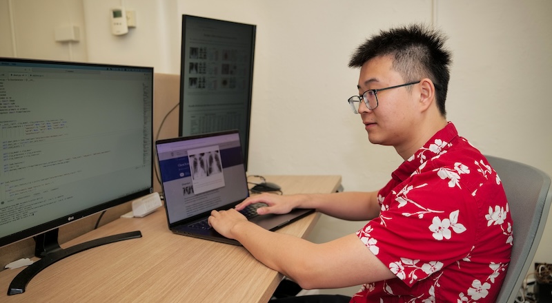 Yue Yang sits at a computer while wearing a Hawaiian shirt.